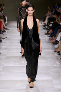 Model walks the runway in a black sequined jumpsuit with a deep V neckline and a velvet cape.