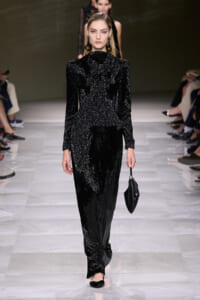 Model walks a runway in a black glitter jumpsuit with structured shoulders, carrying a small black clutch.