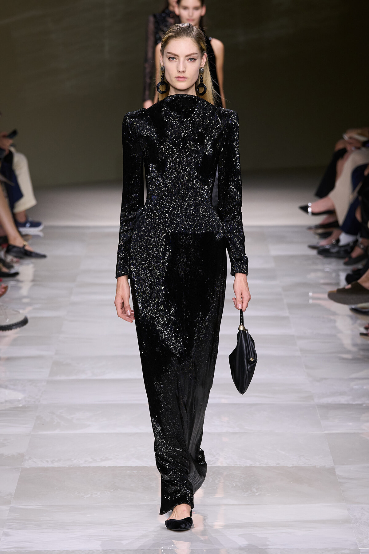 Model walks a runway in a black glitter jumpsuit with structured shoulders, carrying a small black clutch.