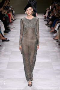 Model on a runway wearing a sheer black-dotted full-length dress with long sleeves and a black beret.