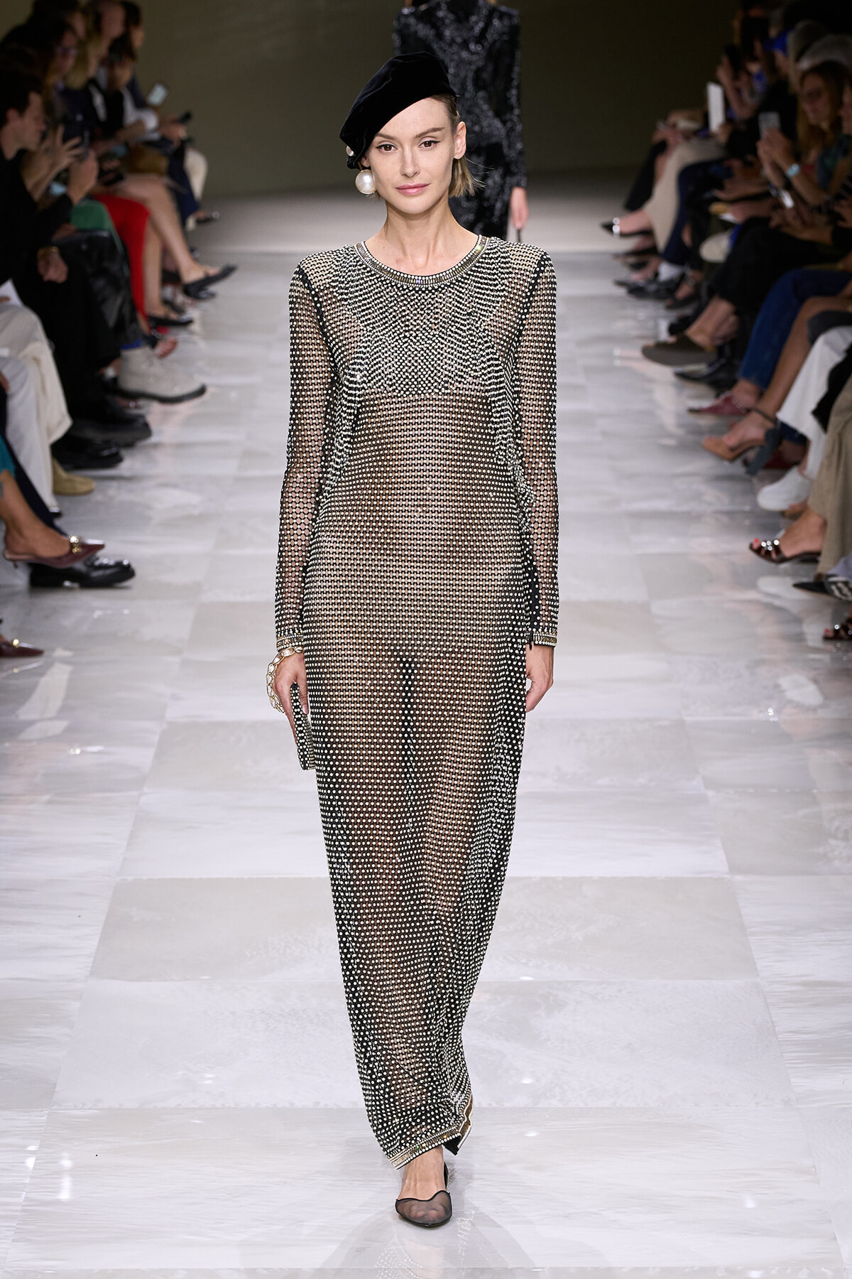 Model on a runway wearing a sheer black-dotted full-length dress with long sleeves and a black beret.