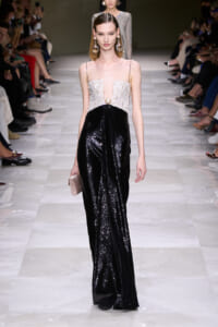 Model walks the runway in a two-tone gown: white beaded bodice with a plunging neckline and a black sequined skirt, clutch in hand.