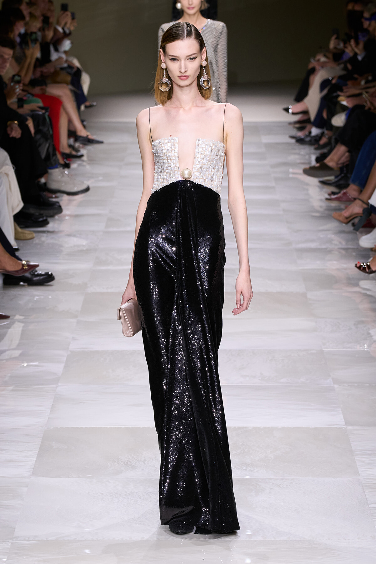 Model walks the runway in a two-tone gown: white beaded bodice with a plunging neckline and a black sequined skirt, clutch in hand.