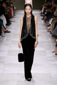 Model walks runway in a black sequin-embellished vest over a sheer top, paired with wide-leg velvet pants and a small black handbag, accessorized with pearl earrings and chunky bracelets.