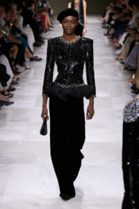 Runway model walking in a black sequined top with pearl detailing, paired with wide-leg velvet pants and a black turban, holding a small black clutch.