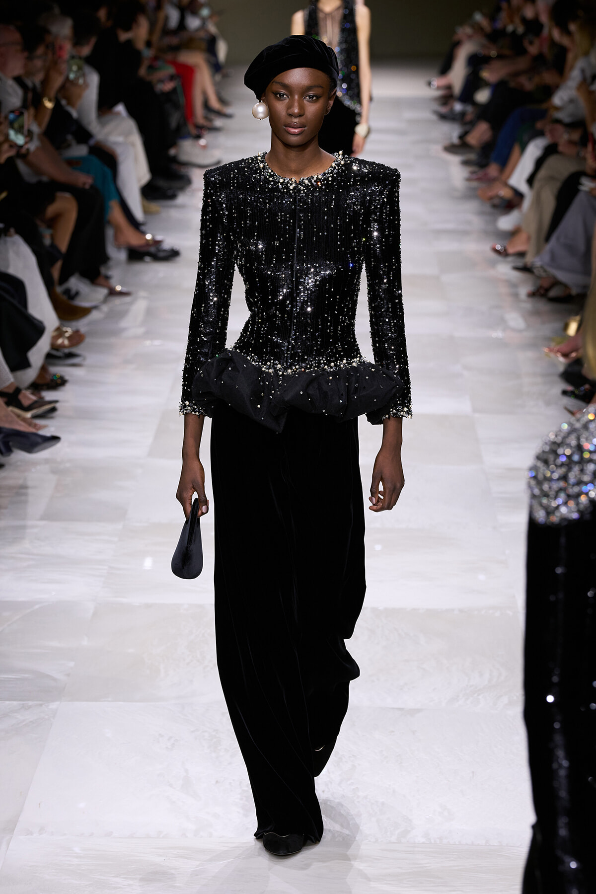 Runway model walking in a black sequined top with pearl detailing, paired with wide-leg velvet pants and a black turban, holding a small black clutch.