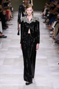 Model walks a black sequined suit with silver embroidered shoulders on a runway, audience flanking the catwalk.