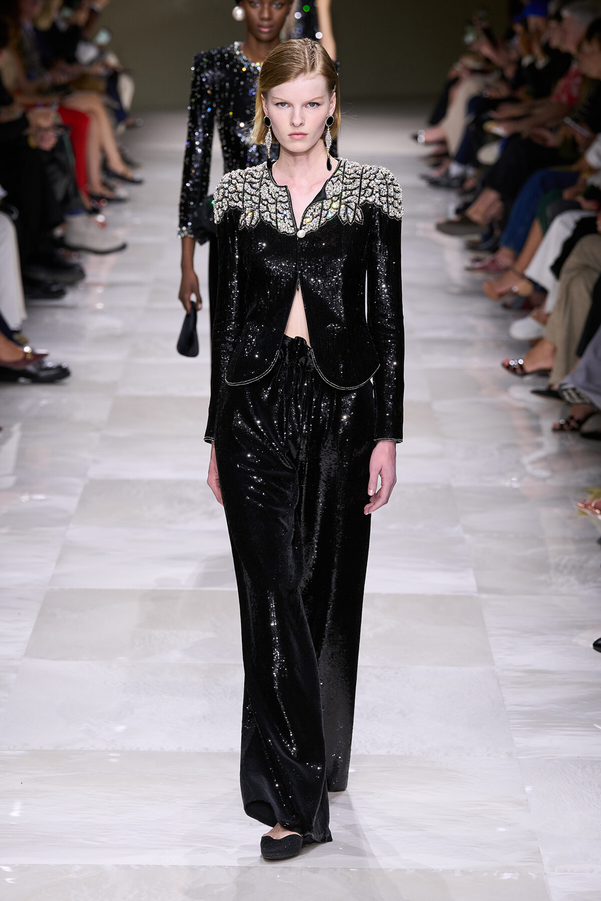 Model walks a black sequined suit with silver embroidered shoulders on a runway, audience flanking the catwalk.