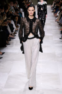 Model walking a fashion runway in a sheer black beaded fringed blouse paired with wide-leg silver sequined pants and black flats.