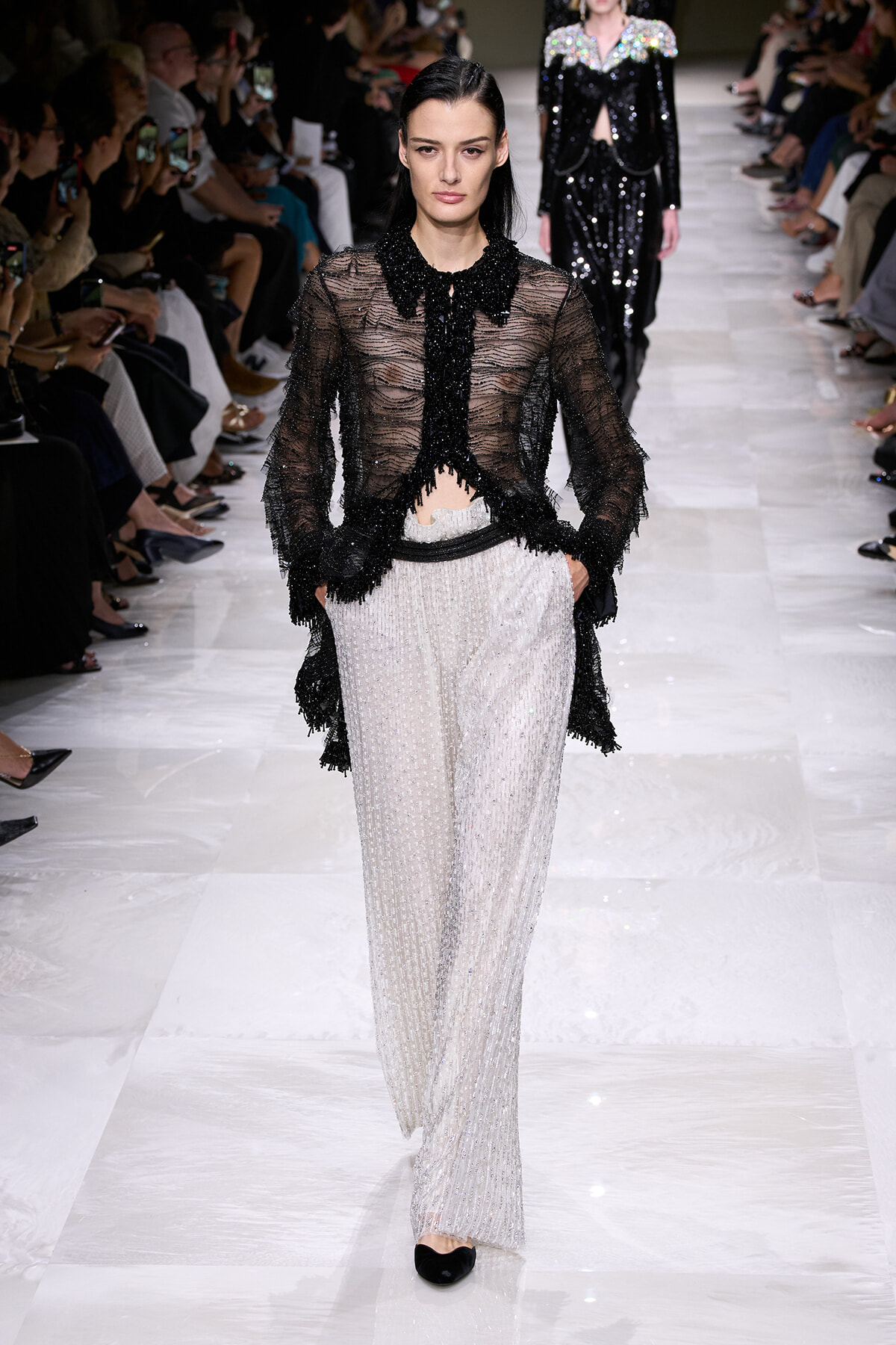 Model walking a fashion runway in a sheer black beaded fringed blouse paired with wide-leg silver sequined pants and black flats.