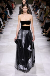 Model walks a runway in a black strapless top and a layered black-and-gray tulle skirt at a fashion show.