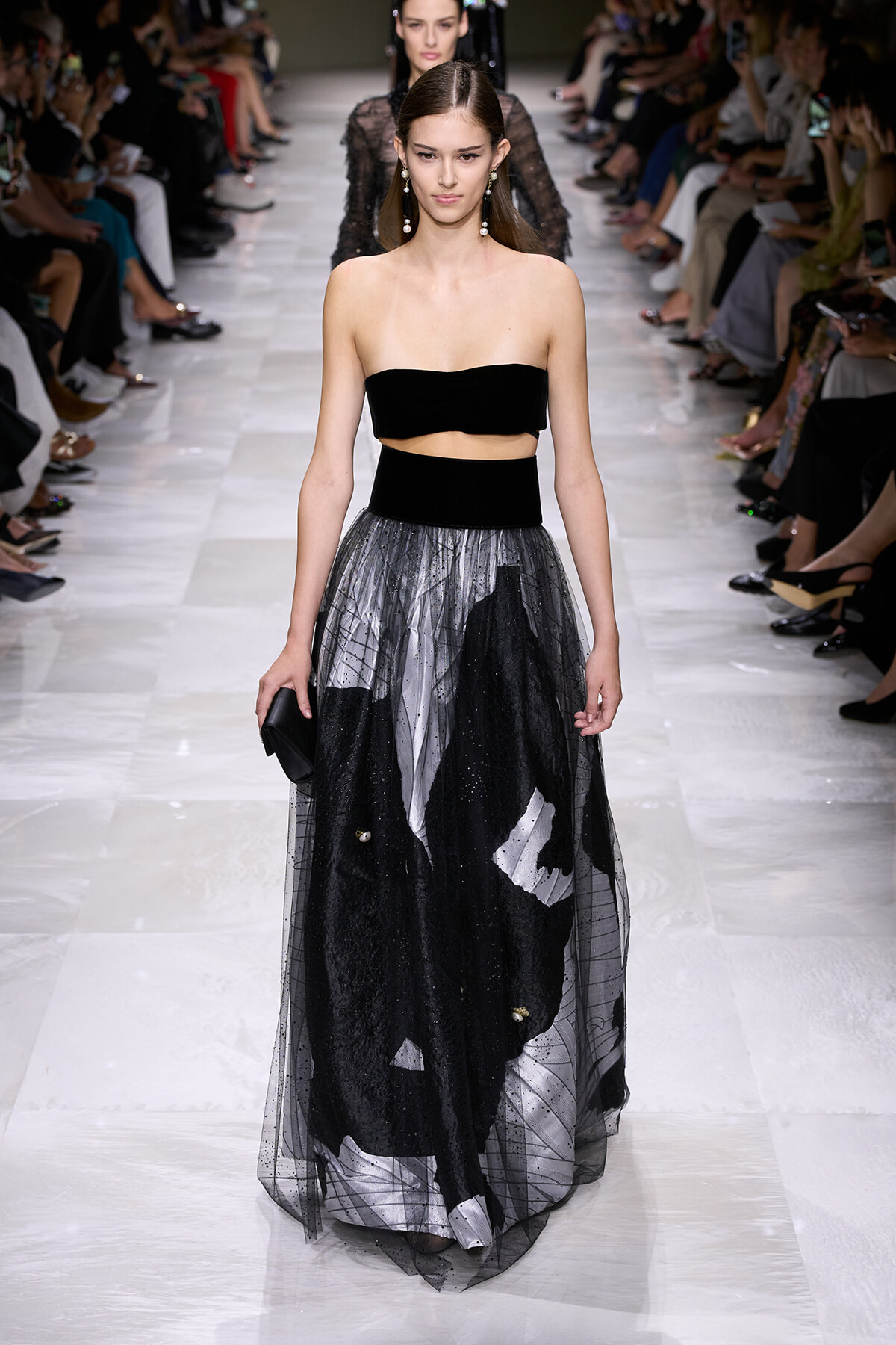 Model walks a runway in a black strapless top and a layered black-and-gray tulle skirt at a fashion show.