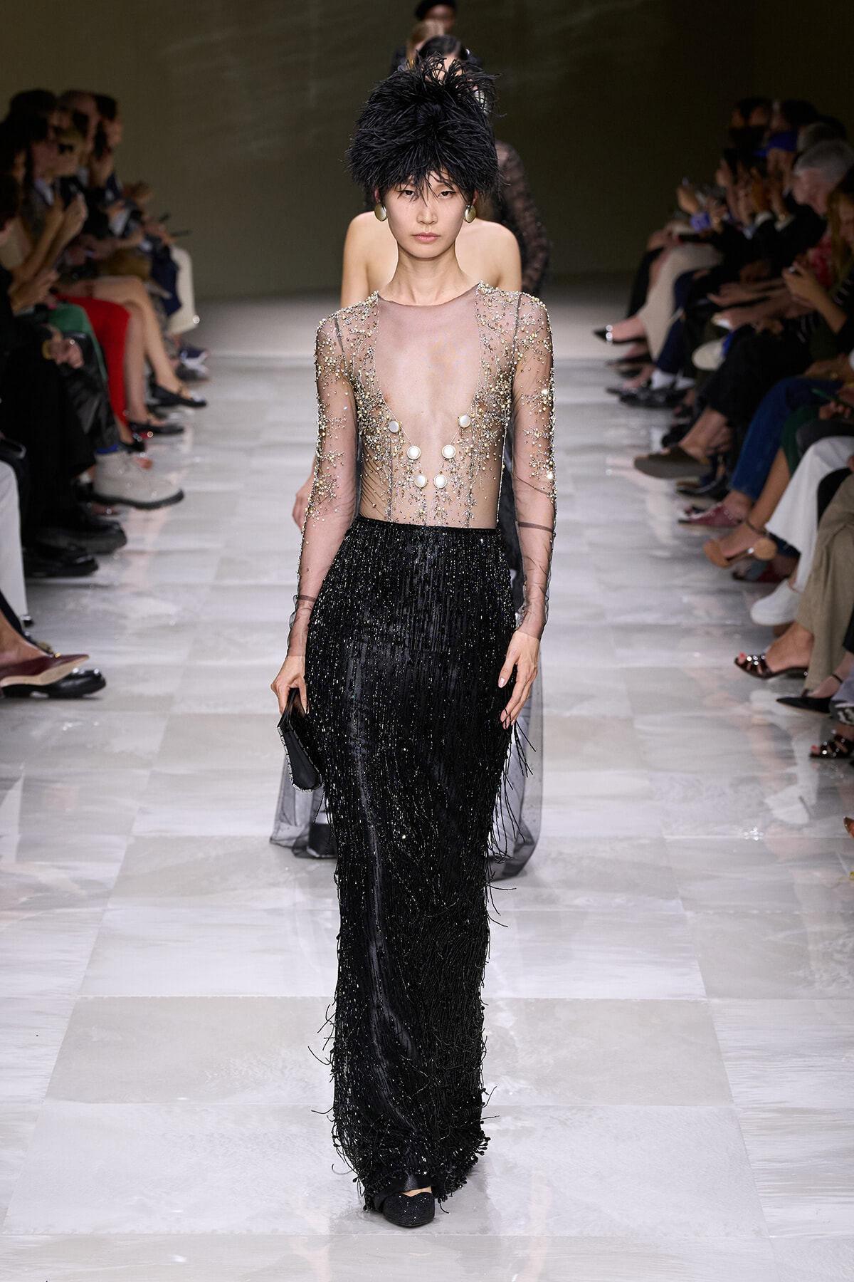 Model on a runway in a sheer beaded top and long black beaded fringe skirt, with a dramatic feathered hat and a small clutch.