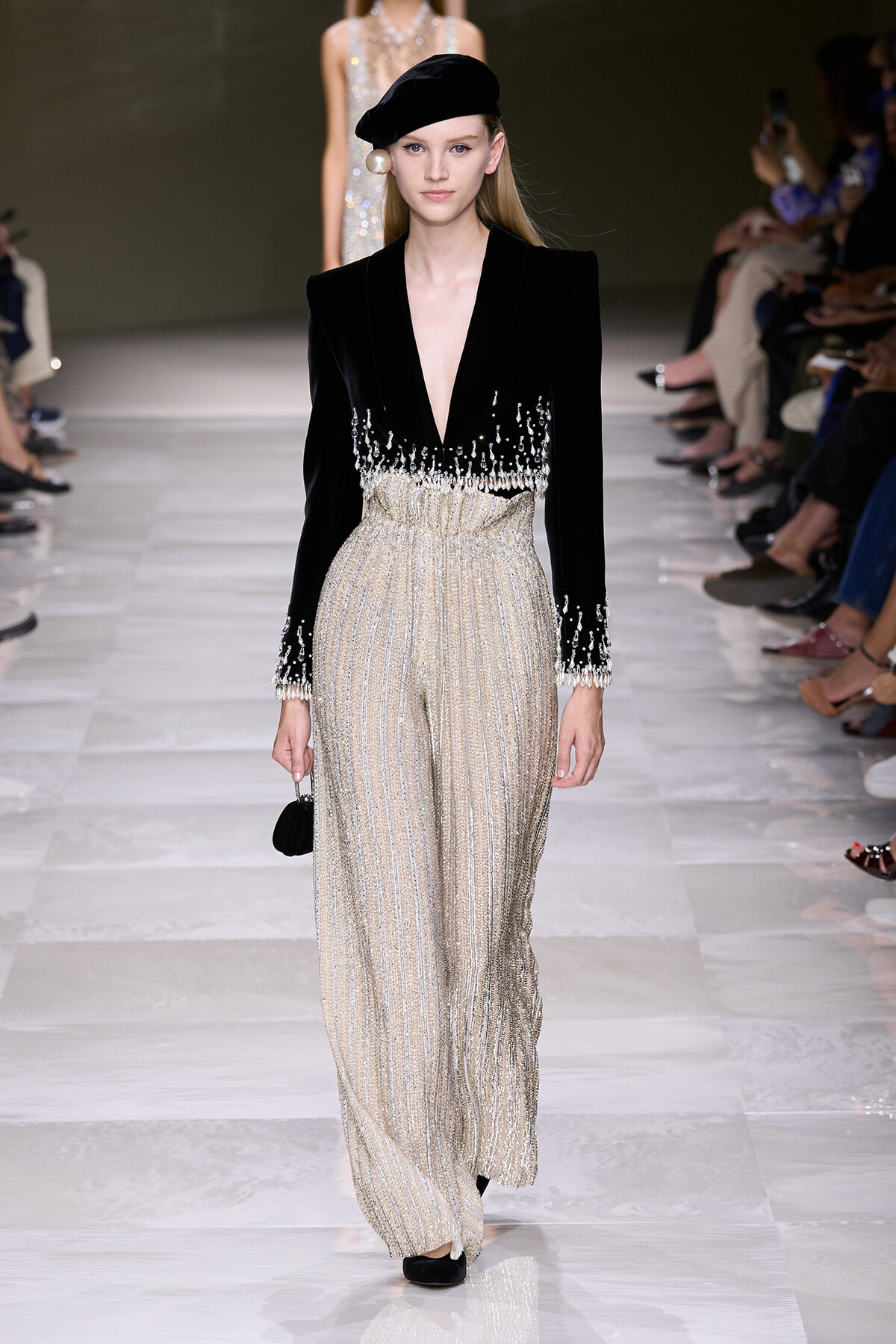 Model on a runway wearing a black velvet deep-V blazer with pearl fringe and sparkling wide-leg metallic striped pants, black beret and small black clutch.