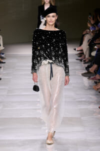 Model on runway wearing a black beaded top with a pearl embellishment gradient into sheer pearl-embellished pants, black beret, and small black handbag.