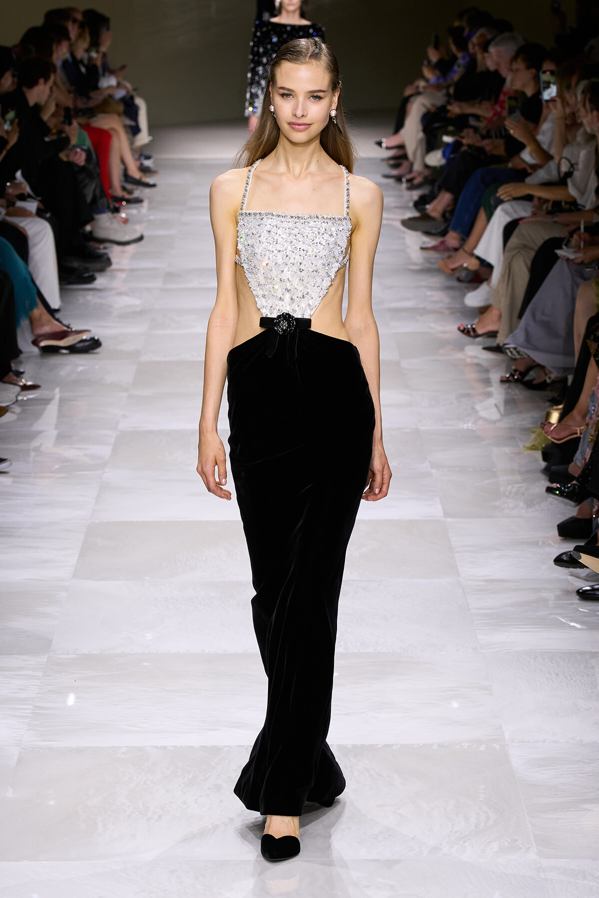 Model on a fashion runway wearing a silver beaded crop top and a high-waisted black velvet gown with a bow at the waist.