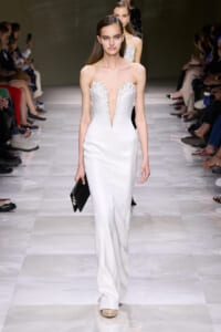 Model walks down a white-runway in a strapless white gown with beaded, plunging neckline, holding a black clutch. Audience seated on both sides.