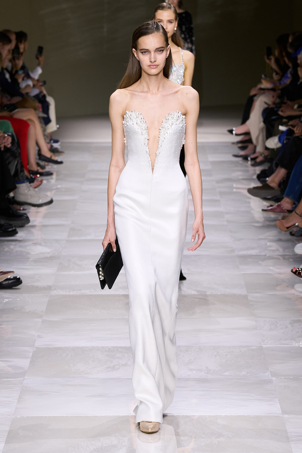 Model walks down a white-runway in a strapless white gown with beaded, plunging neckline, holding a black clutch. Audience seated on both sides.