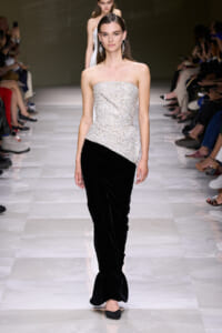Model on a runway wearing a strapless silver beaded top with an asymmetrical peplum over black velvet wide-leg pants, accessorized with large dangling earrings.