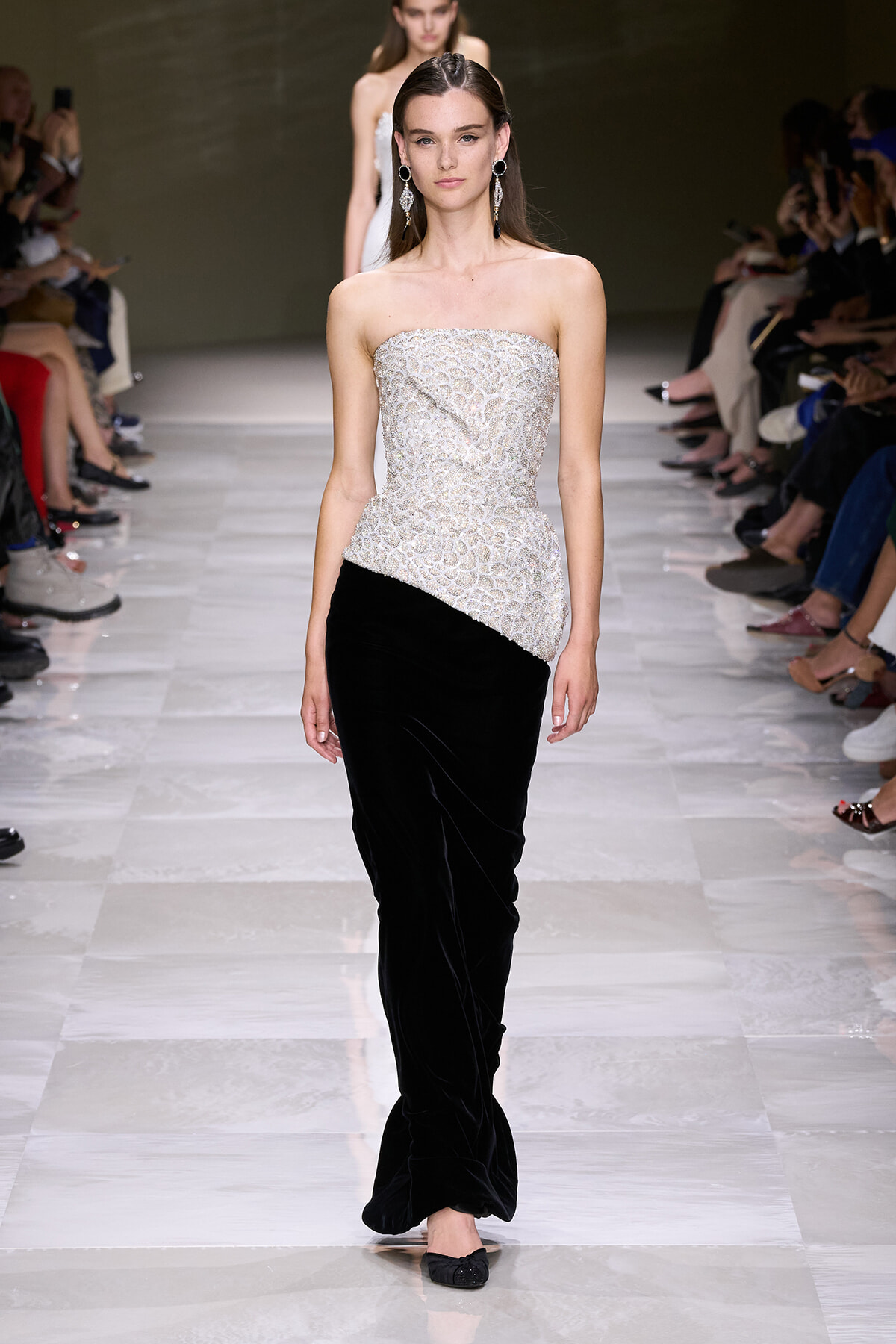 Model on a runway wearing a strapless silver beaded top with an asymmetrical peplum over black velvet wide-leg pants, accessorized with large dangling earrings.