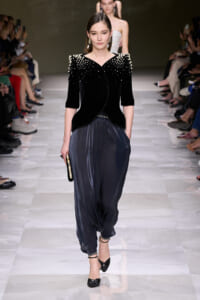Model walks a runway in a black velvet top with pearl embellishments and loose navy pants, holding a small clutch.