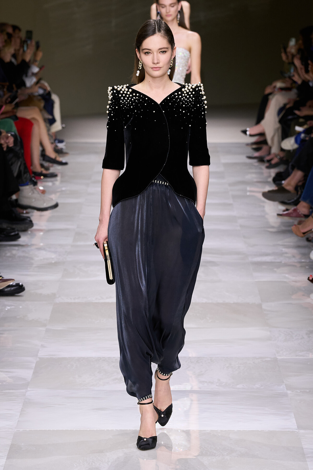 Model walks a runway in a black velvet top with pearl embellishments and loose navy pants, holding a small clutch.