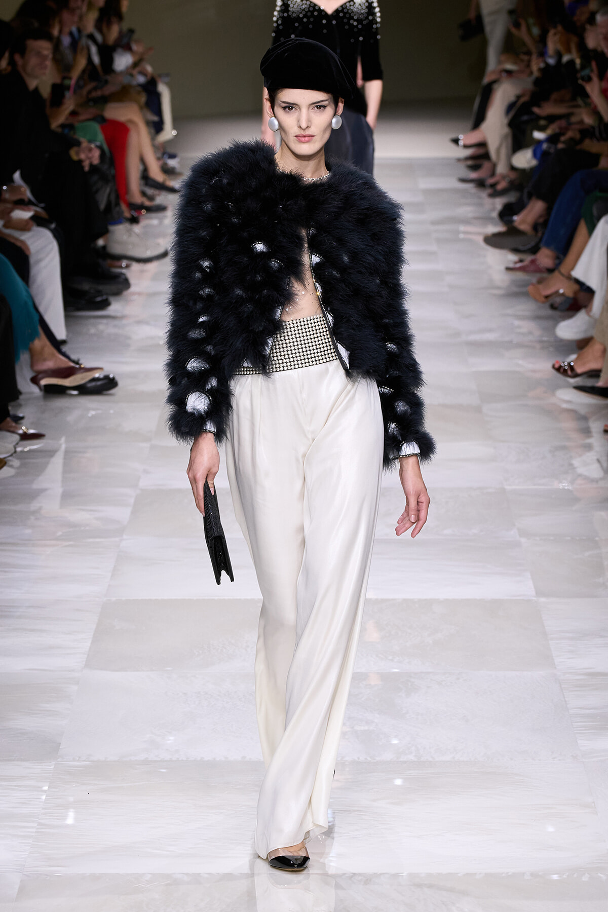 Model on a runway wearing a black furry cropped jacket, white wide-leg satin pants, and a studded belt, carrying a black clutch and wearing a black beret.