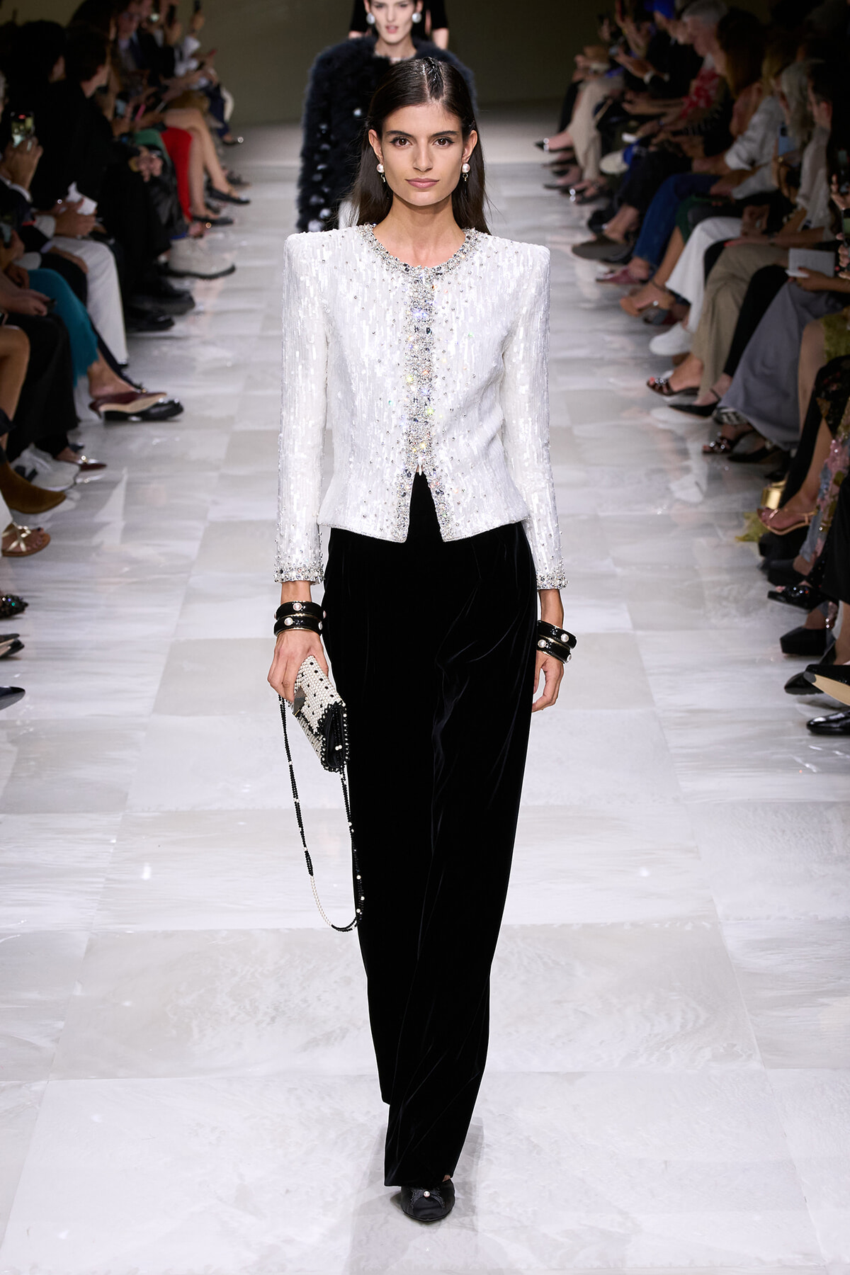 Model walks the runway in a white sequined blazer with crystal trim and black velvet wide-leg pants, carrying a beaded clutch with a chain strap.