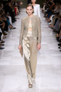 Model walking the runway in a beige embellished jacket with matching sheer top and metallic champagne wide-leg pants, carrying a beige clutch at her side.