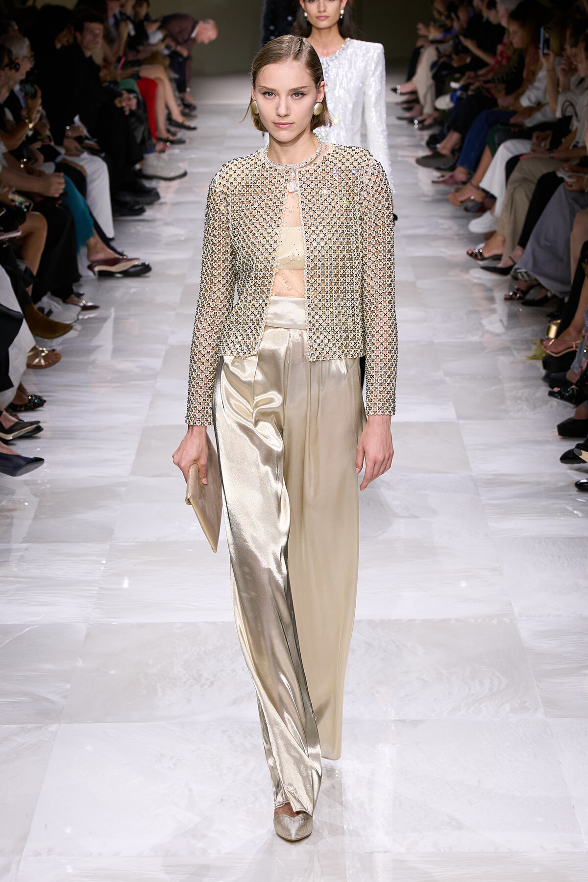 Model walking the runway in a beige embellished jacket with matching sheer top and metallic champagne wide-leg pants, carrying a beige clutch at her side.