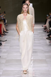Model walks the runway in a cream pearl-embellished cape over a shimmering ivory jumpsuit, posture poised and hands in pockets.