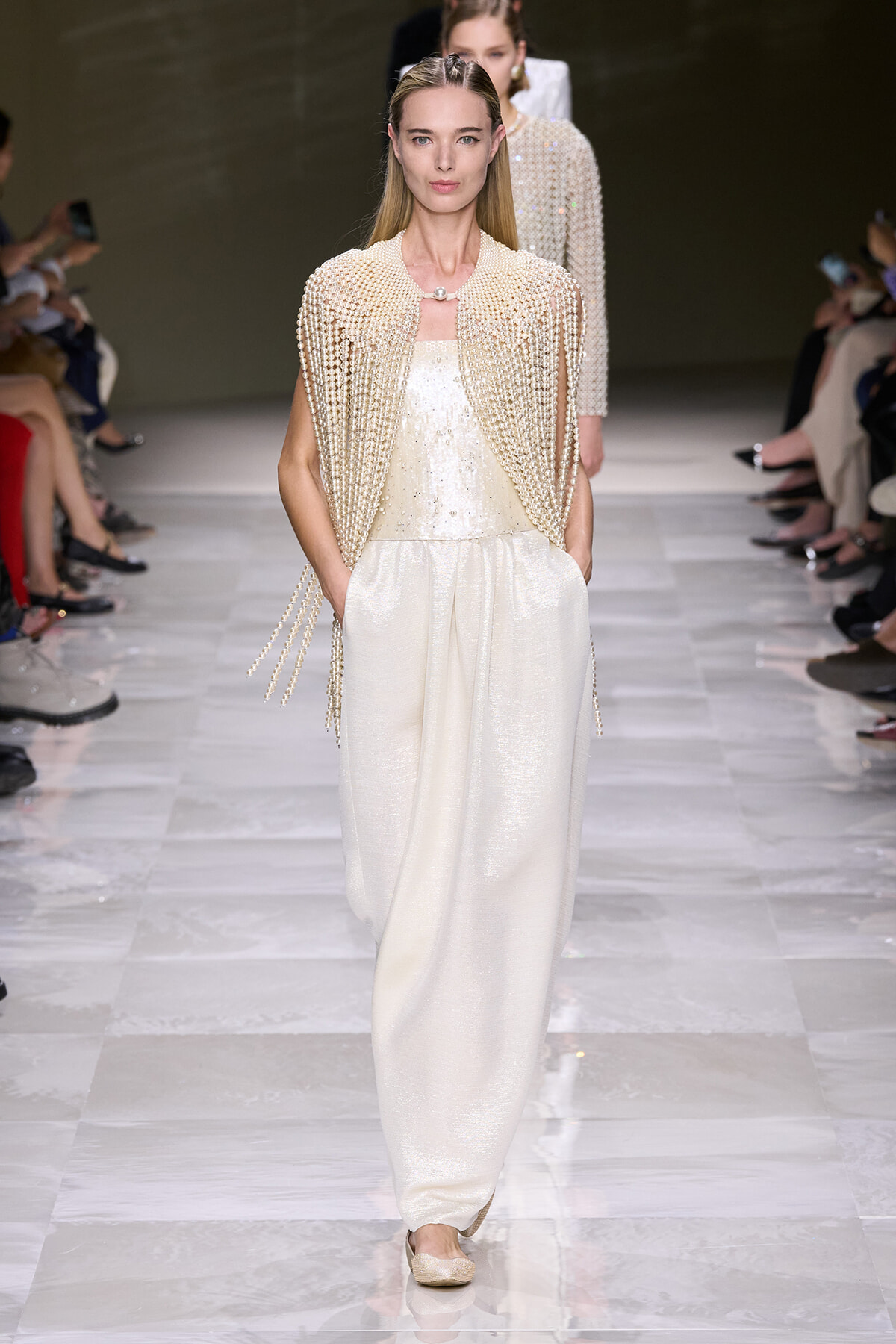Model walks the runway in a cream pearl-embellished cape over a shimmering ivory jumpsuit, posture poised and hands in pockets.