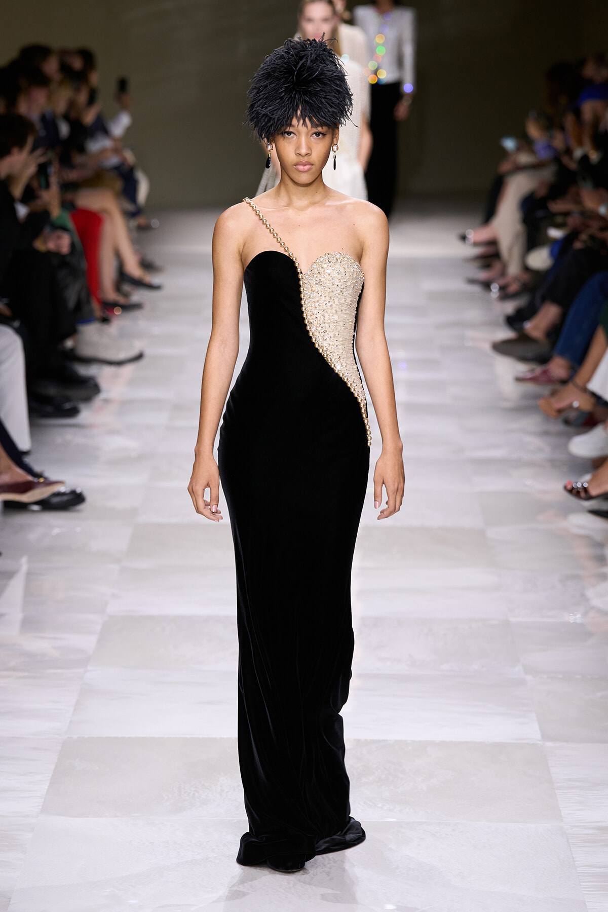 Model walking the runway in a black velvet, one-shoulder gown with a gold beaded side panel and a black feathered headpiece.