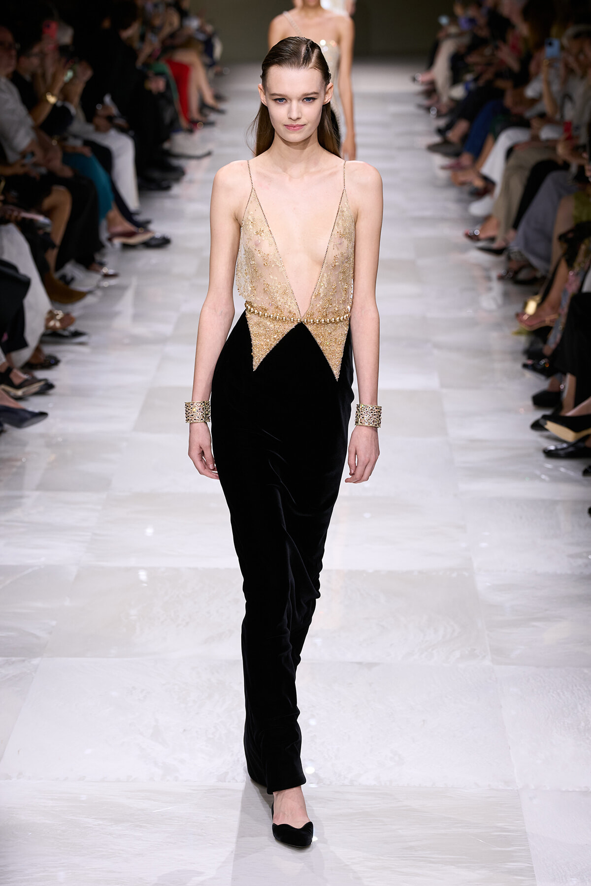 Model walking a runway in a gold-beaded sheer plunging top and black velvet pants, with matching gold cuffs on both wrists.