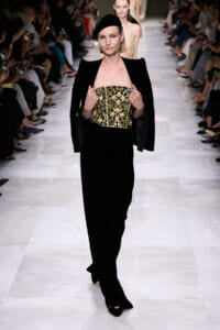 Model walks the runway in a black velvet outfit with a gold embellished strapless bodice and dramatic shoulders, accessorized with a black headband and pearl earring.