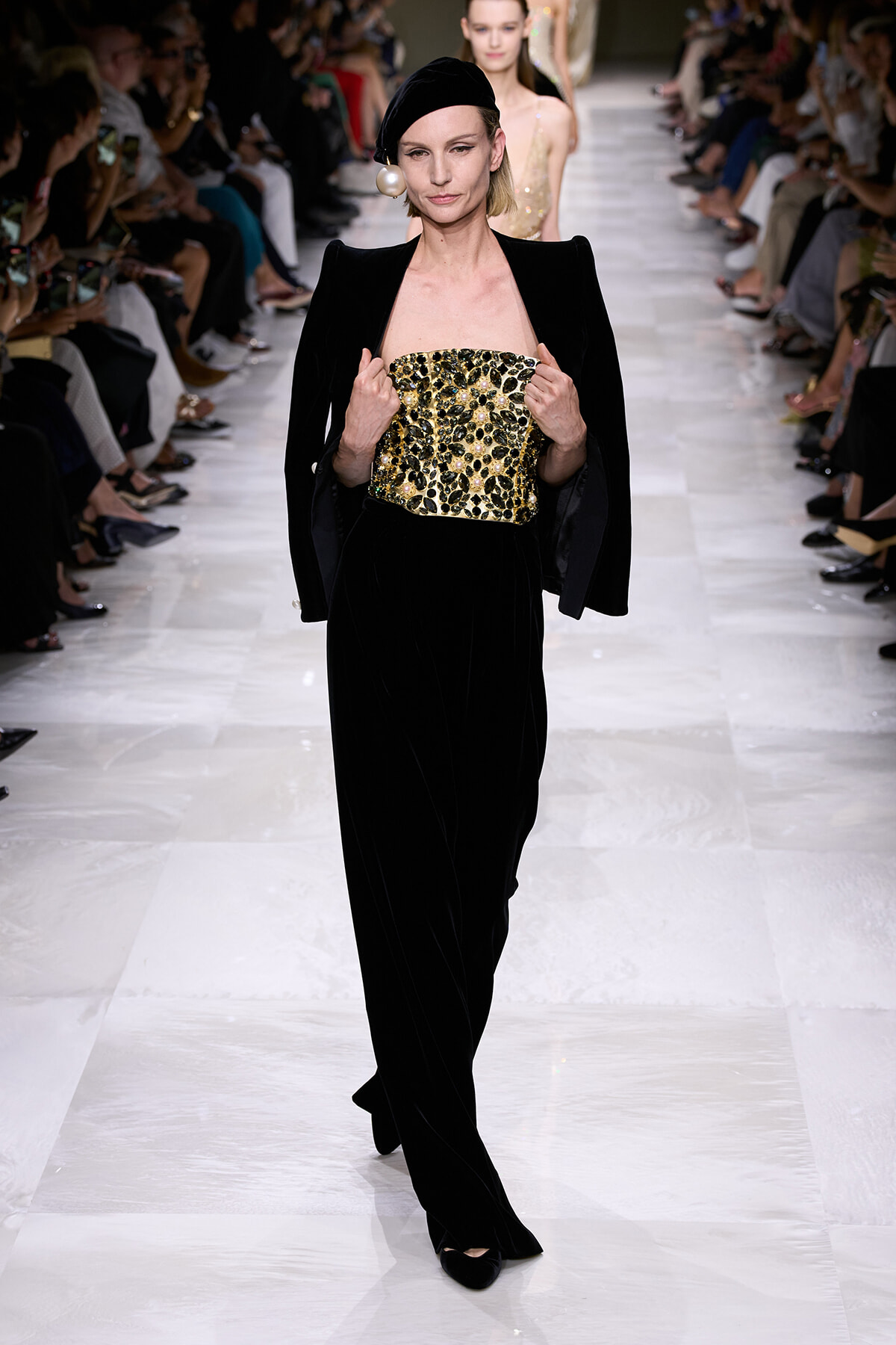 Model walks the runway in a black velvet outfit with a gold embellished strapless bodice and dramatic shoulders, accessorized with a black headband and pearl earring.