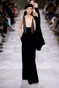 Model walks a fashion runway in a black velvet jumpsuit with deep V neckline and beaded suspenders, paired with a black beret and oversized pearl earring, while carrying a matching coat over her shoulder.