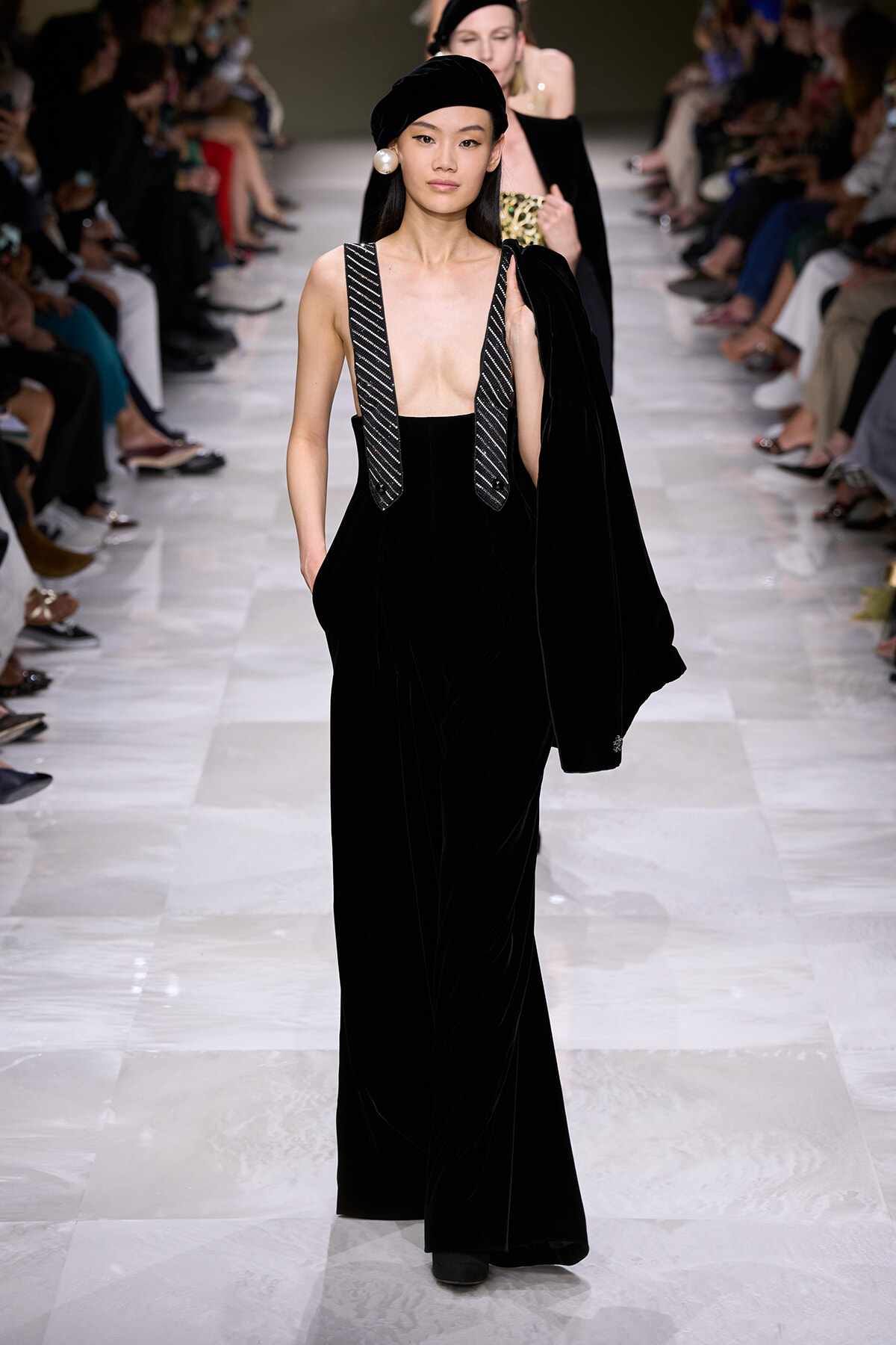 Model walks a fashion runway in a black velvet jumpsuit with deep V neckline and beaded suspenders, paired with a black beret and oversized pearl earring, while carrying a matching coat over her shoulder.