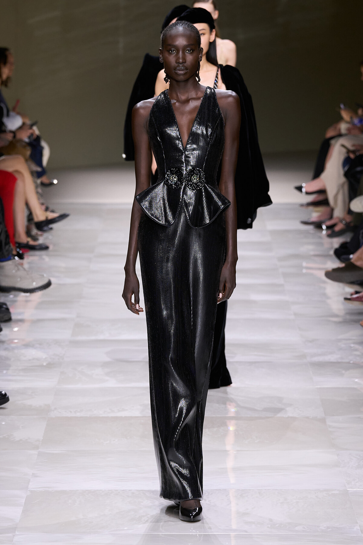 Model walking the runway in a shiny black metallic jumpsuit with a plunging V-neck and peplum waist accented by a jeweled belt on the hips.