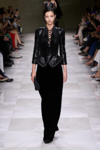Model walks a black sequined blazer over dark velvet wide-leg pants on a runway, accessorized with a bold beaded necklace.