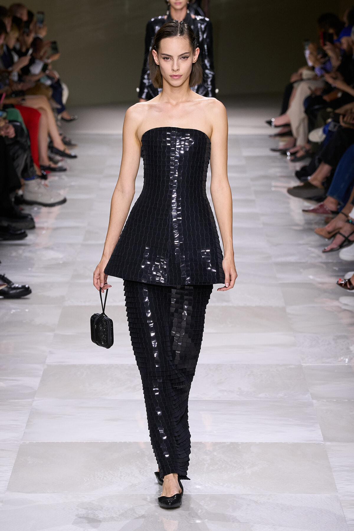 Model walks the runway in a strapless, glossy black two-piece outfit with a square-textured pattern and a small black clutch on the left hand.