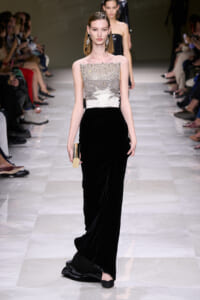 Model walks a runway in a strapless silver-to-black velvet gown with chandelier earrings and a gold clutch on the left hand.