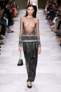 Model walks a runway in a sheer, beaded top and long black fringe pants, carrying a small pearl-embellished black handbag; audience sits on both sides.