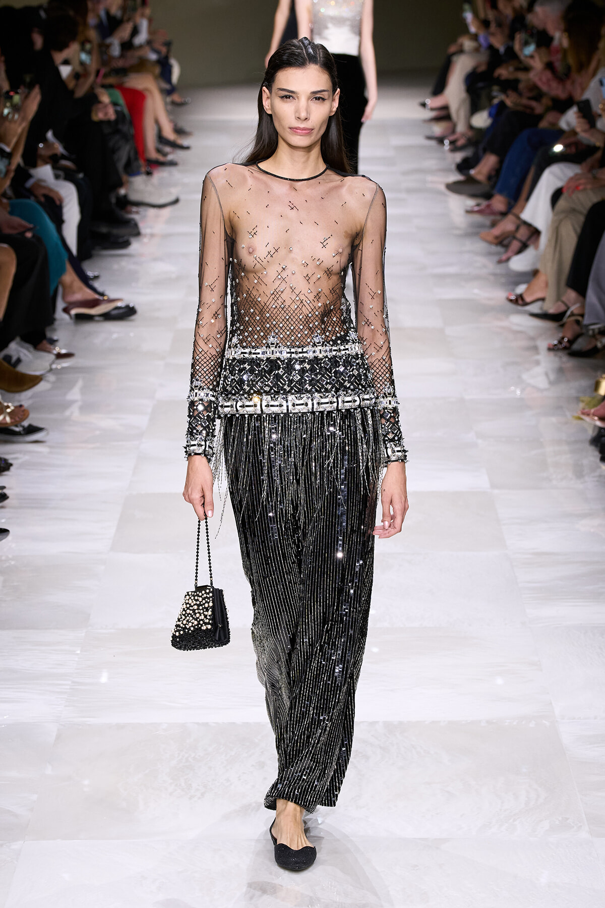 Model walks a runway in a sheer, beaded top and long black fringe pants, carrying a small pearl-embellished black handbag; audience sits on both sides.