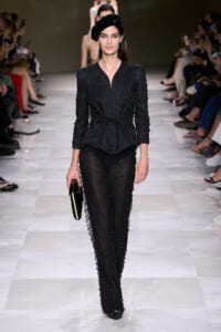 Model walks the runway in a black beaded two-piece suit with a knotted waist, sheer beaded pants, a black beret, and a small black clutch.
