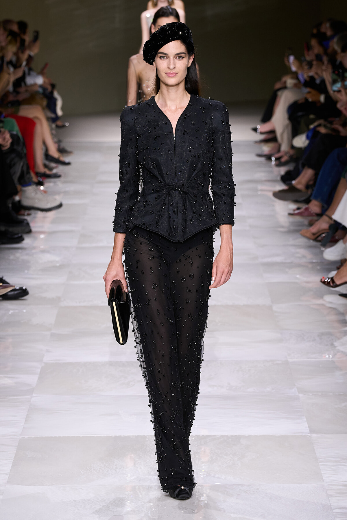 Model walks the runway in a black beaded two-piece suit with a knotted waist, sheer beaded pants, a black beret, and a small black clutch.