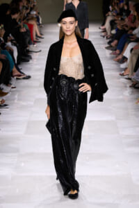 Model on a runway wearing a black velvet blazer, sheer beaded top, and sparkling wide-leg sequined pants with a black beret.