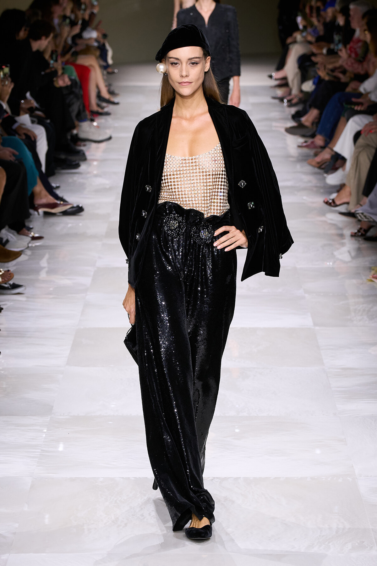 Model on a runway wearing a black velvet blazer, sheer beaded top, and sparkling wide-leg sequined pants with a black beret.
