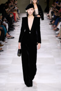 Model walks the runway in a black velvet pantsuit with a deep plunging neckline, beret, and pearl earrings, carrying a small black handbag.