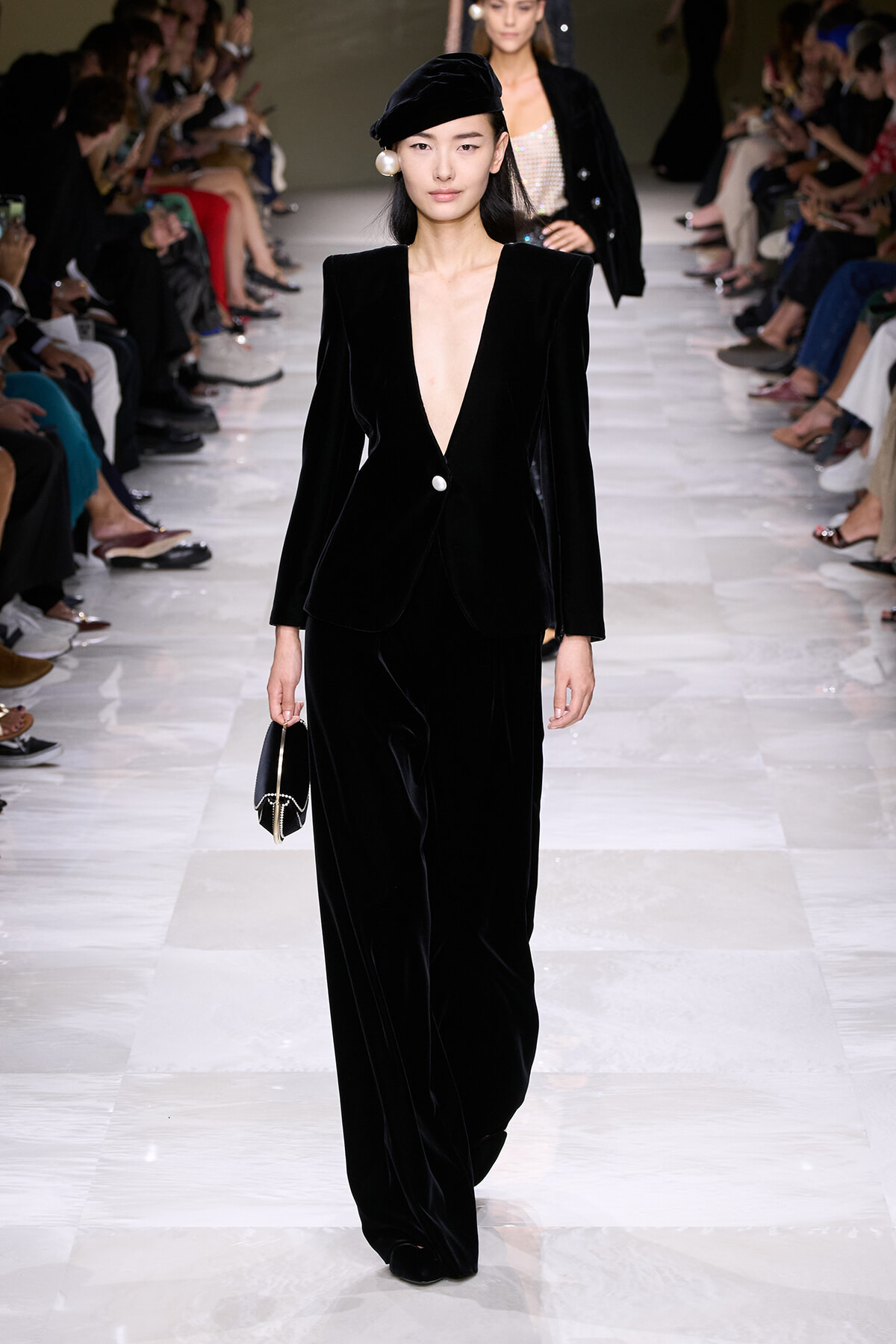 Model walks the runway in a black velvet pantsuit with a deep plunging neckline, beret, and pearl earrings, carrying a small black handbag.